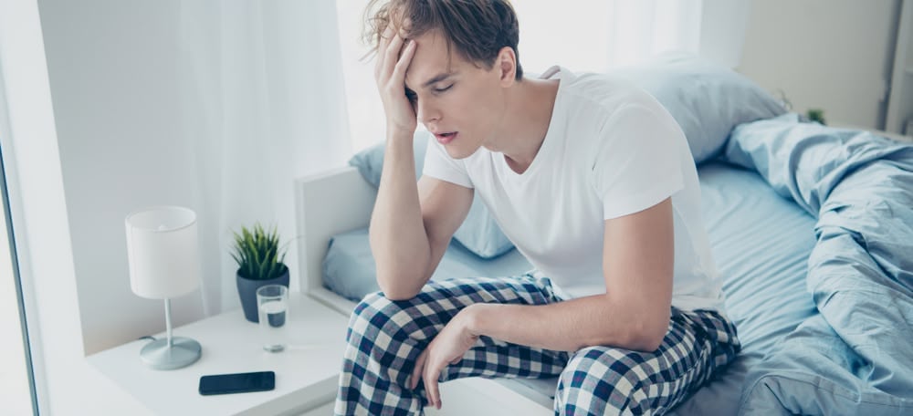Portrait of exhausted unhappy man after hangover party have terrible fatigue migraine catch cold suffer sit on bed wear checkered plaid pajama in house indoors