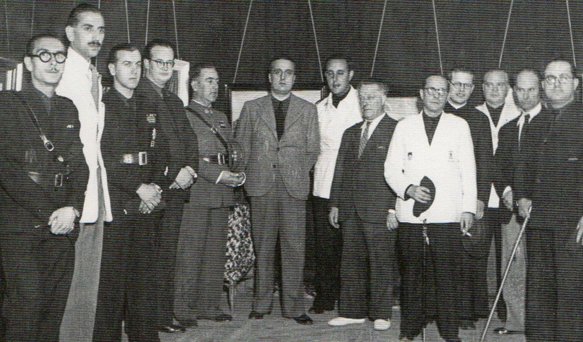 a plana major, d'esquerra a dreta: Francesc Truyols, Joaquim Garriga, Mateu Abella, Andreu Flaqué, ?, Josep Mª Marcet (al centre), Ramon Grau, Lluís Mas, Miquel Sala Viñals, mossèn Ernest Mateu, Vila Puig, Pau Mª Llonch Gambús (tinent d'alcalde de Cultura) . Exposició dels cartells de la Festa Major a Belles Arts (31 de juliol de 1941)